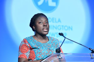 2014 Fellow Adepeju Jaiyeoba delivers a speech at a podium at the 2019 Mandela Washington Fellowship Summit after she is presented with the inaugural Leadership Impact Award. She has her hair pulled back and is wearing an orange and teal patterned dress.