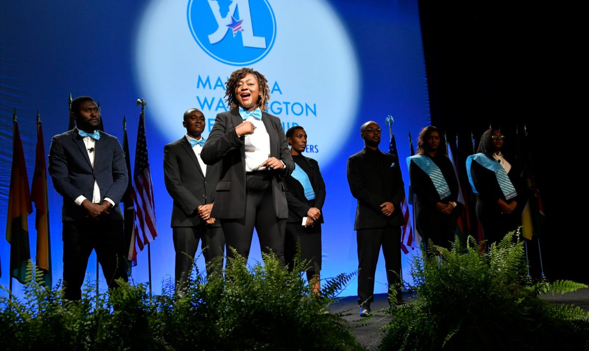 2017 Alumna Ruzivo Chonyera speaks at the 2018 Mandela Washington Fellowship Summit Closing Session. She wears a black suit, light blue tie, and natural hair and stands in front of a line of other alumni in similar dress.