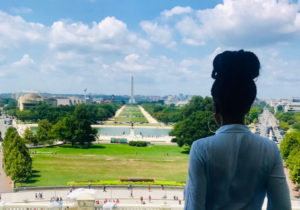2019 PDE Fellow Balkissa Gambo Illo Daoura observes the Washington Monument from the U.S. Capitol. The obelisk rises in the distance, with green grass, trees, and a reflecting pool in the foreground in front of Balkissa, whose back is to the camera.