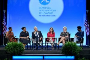 Panelists speak on stage during the 2019 Summit. There are 6 speakers sitting in chairs.