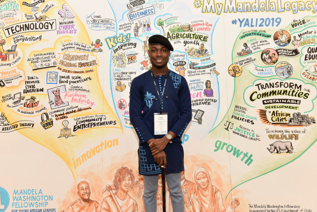 Vital Sounouvou stands in front of a graphic recording depicting pledges from the #MyMandelaLegacy campaign; black man wearing a blue tunic and black beret, holding a cane.