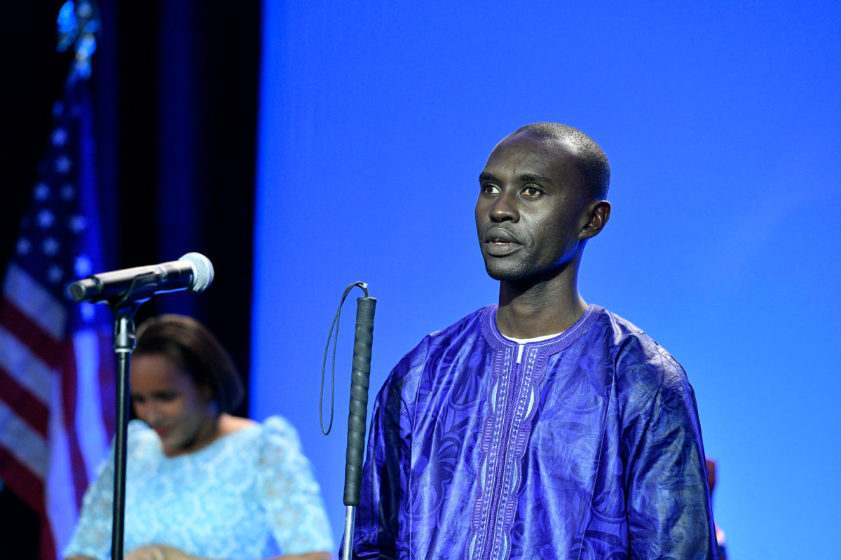 Alieu Jaiteh speaks at a microphone; background is blue; subject holds sight cane.