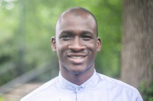 Ibrahima smiles for the camera, standing in nature, wearing a light blue collared shirt.