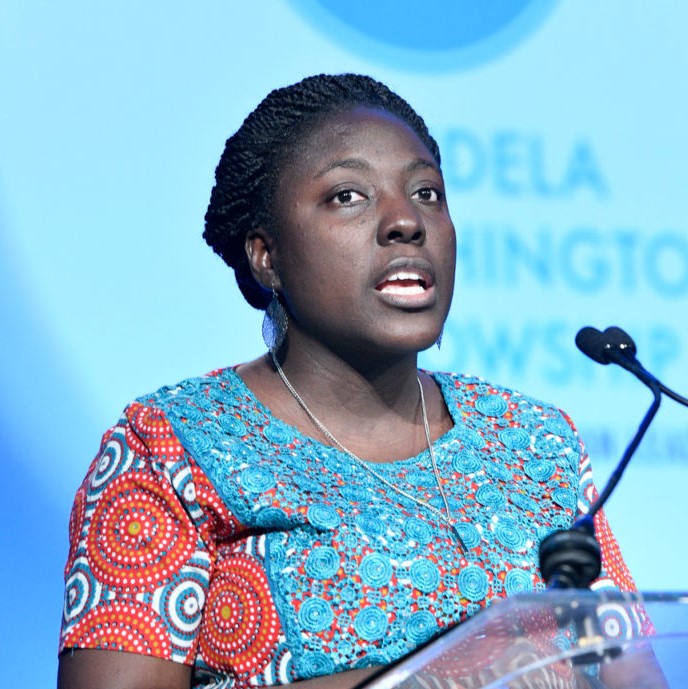 Black woman speaks at a podium. She is wearing an orange and teal patterned dress.