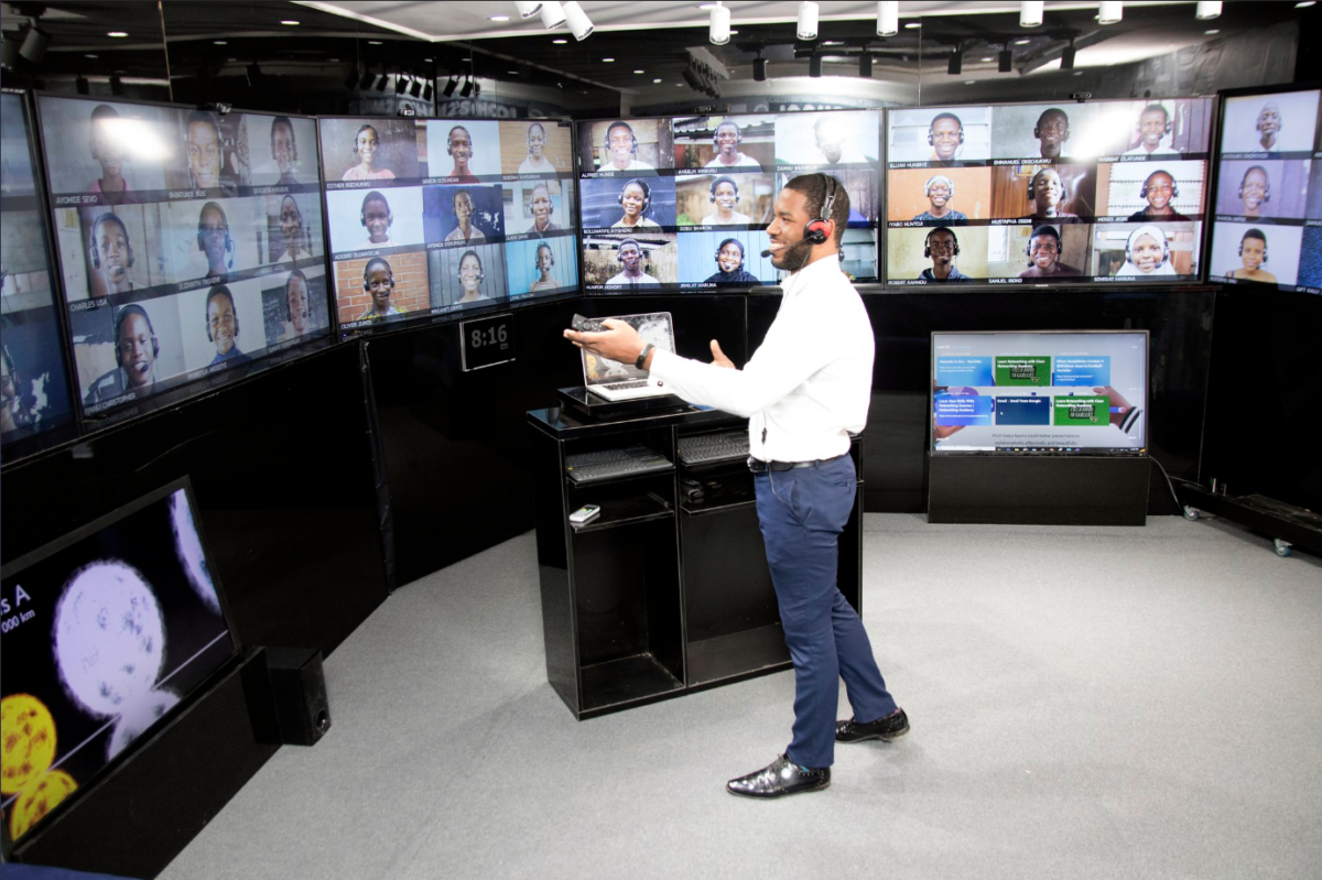 Otto stands in a room surrounded by computer screens showing children joining a virtual class. He is wearing a white shirt, blue pants, and black shoes and a headset, with his arms outstretched.