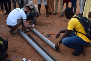 Scott Massey works with a group of men to measure and cut a hollow pole, encircled by a group of bystanders, whose feet are visible.