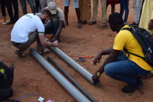 Scott Massey works with a group of men to measure and cut a hollow pole, encircled by a group of bystanders, whose feet are visible.