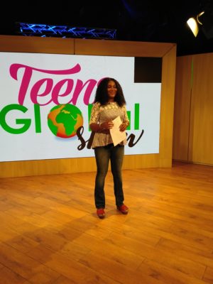 A woman stands on a television set with the words "Teenz Global Show" on a screen behind her.