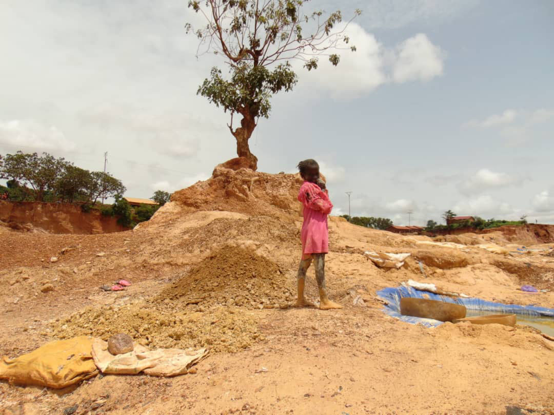 A young girl stands in a sandy area that has been mined. She is surrounded by dirt in a crater in the earth that takes up the whole frame of the photo.