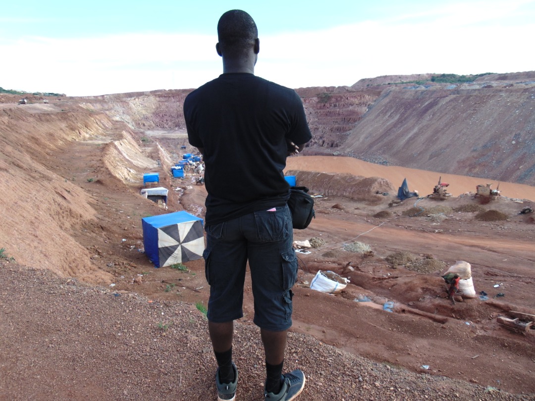 A man looks into the distance at a mine, a large pit with water in it. People work in the vicinity.