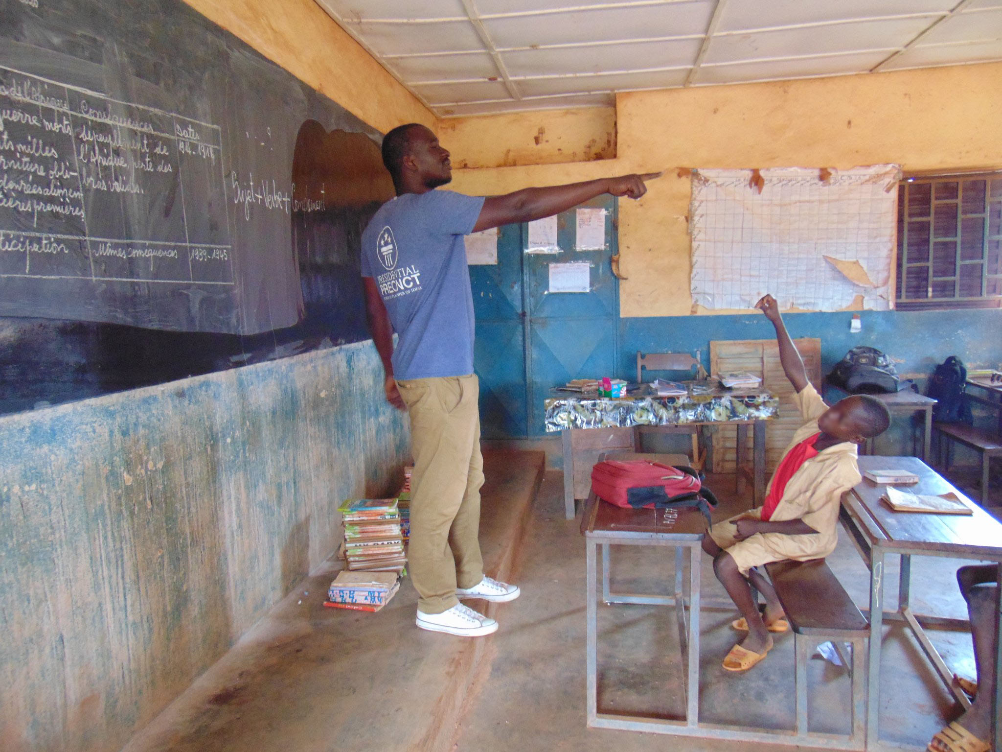 A man stands at the front of a classroom, pointing to someone out of frame. A young boy sits at a desk in front of him with his hand up.