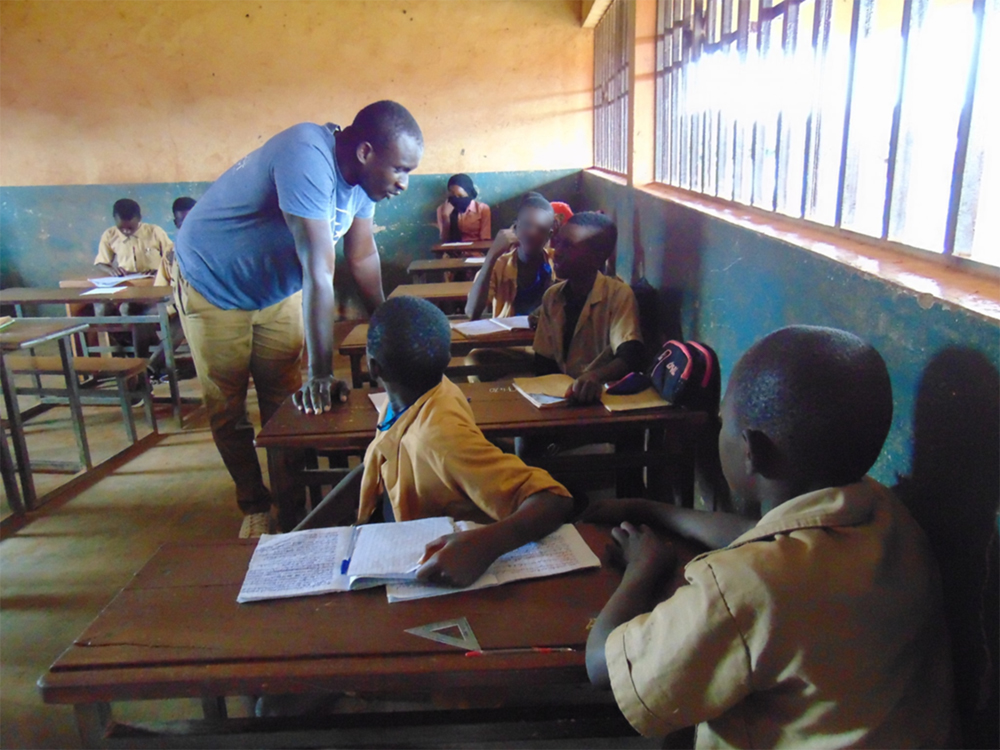 A man speaks with children, seated at desks along the wall of a classroom.
