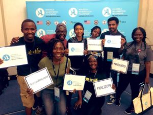 A group of people smile for the camera holding signs featuring the Fellowship logo, #YALI2019, #MyMandelaLegacy.