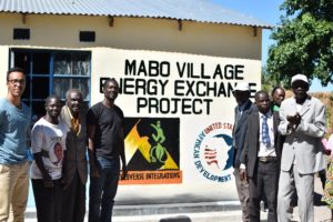 A group of men stand by a sign on a building that says "Mabo Village Energy Exchange Project"