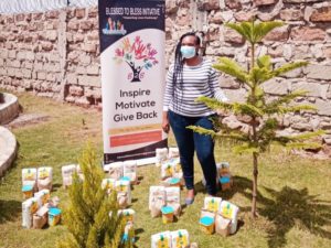 A woman stands outdoors by a sign for the Blessed to Bless Initiative and care packages, next to a small tree.
