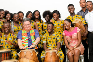 A group of people smiles and plays drums, wearing traditional dress in yellow pattern