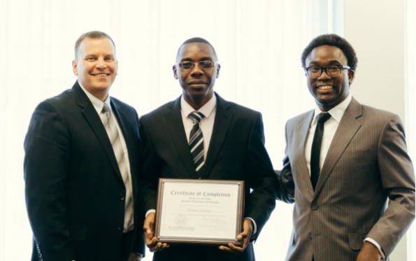 Three men smile at a camera; man in middle holds a certificate of completion