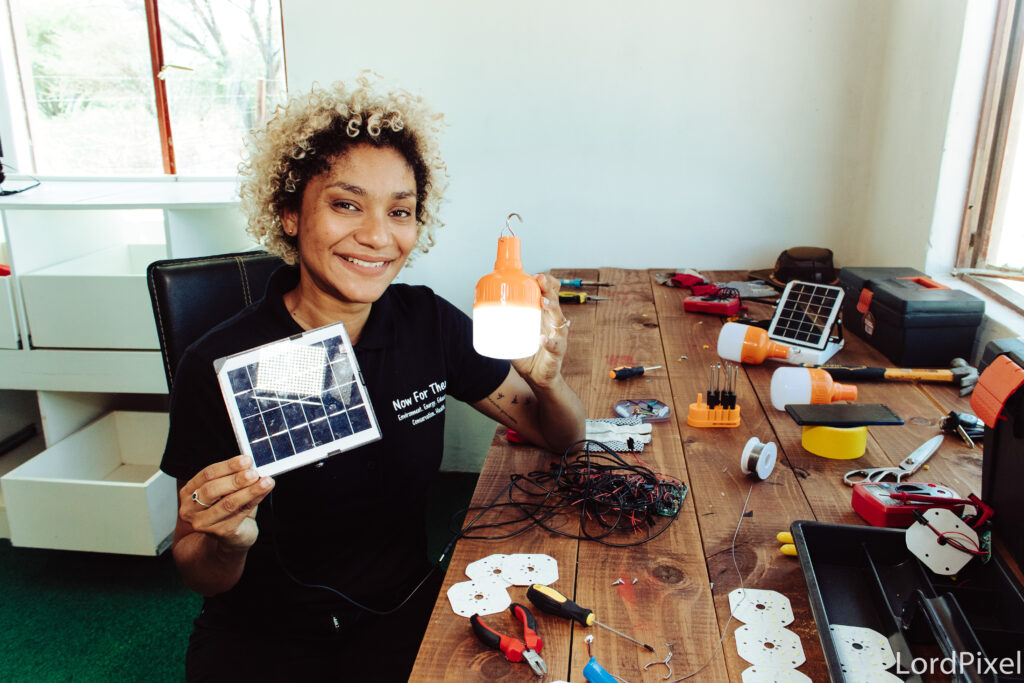 Sarah smiles for the camera and holds a small solar panel and light at a work bench