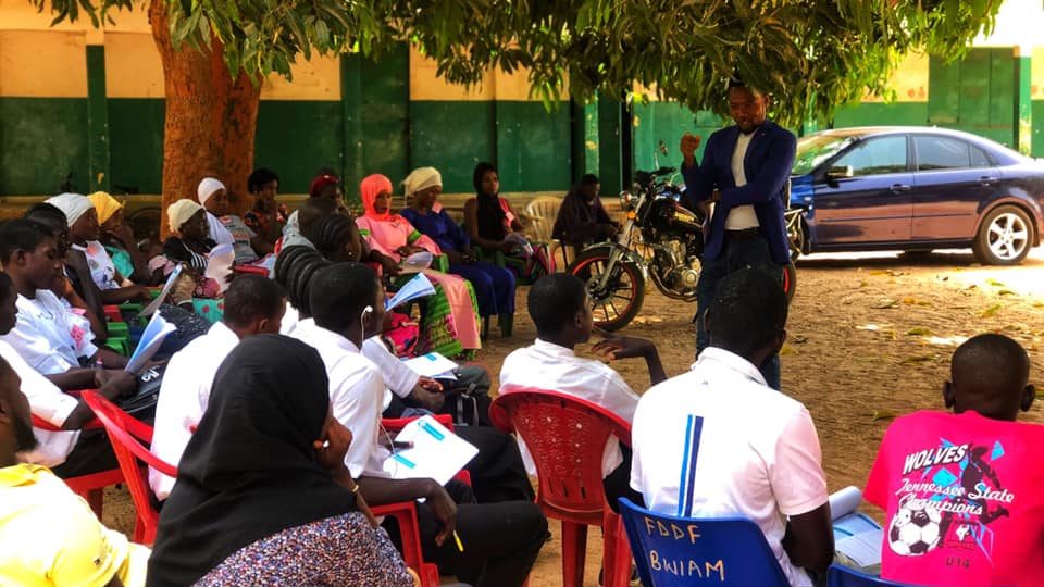 A man speaks before an outdoor workshop event. He is standing under a tree and the audience is in chairs.