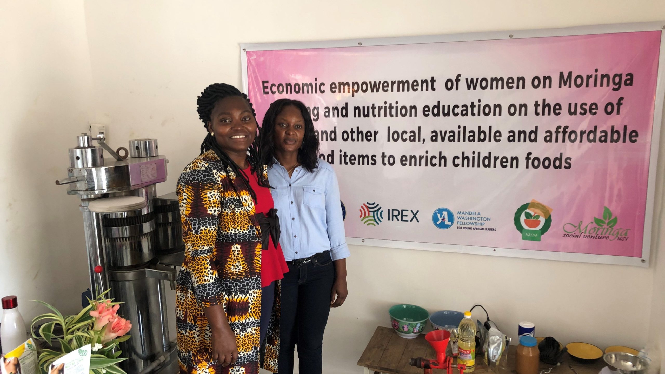 2 women stand in front of a poster on a wall, which says "Economic empowerment of women on moringa farming and nutrition education on the use of moringa and other local, available and affordable food items to enrich children foods."