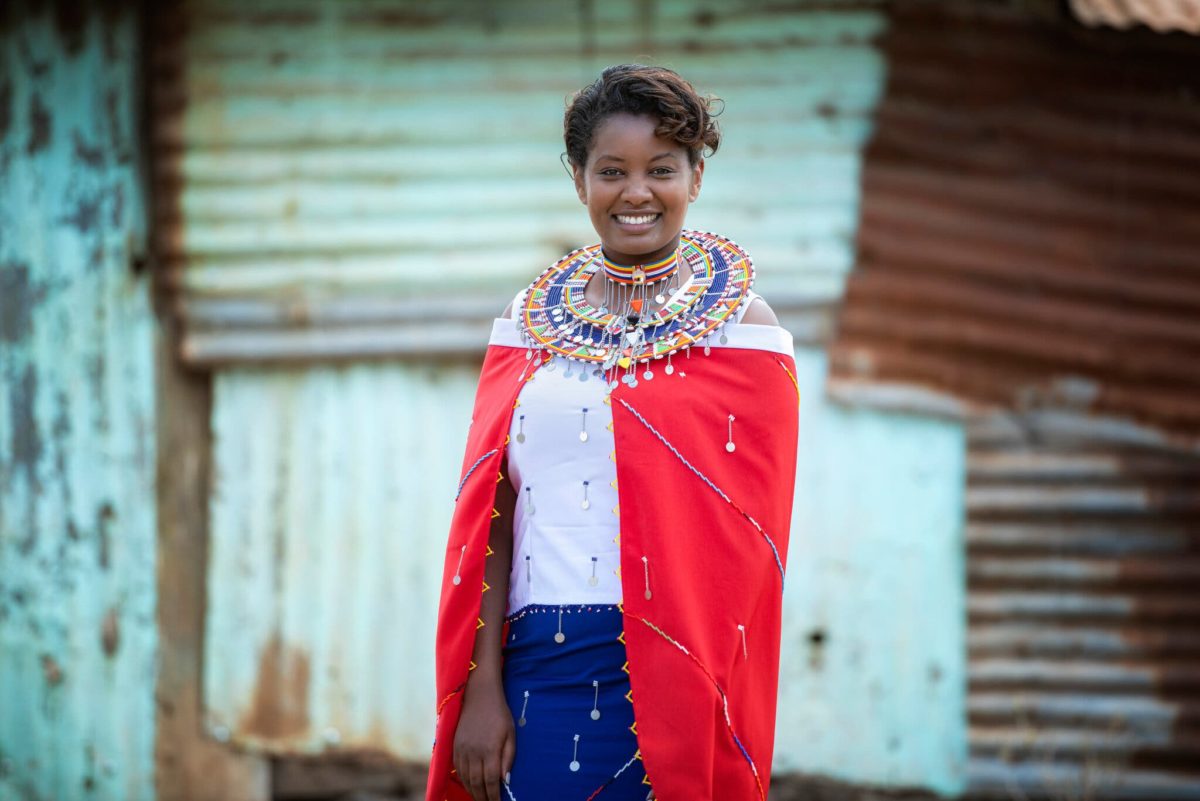 Young woman in traditional Maasai dress and jewelry stands in an open area with a structure behind her