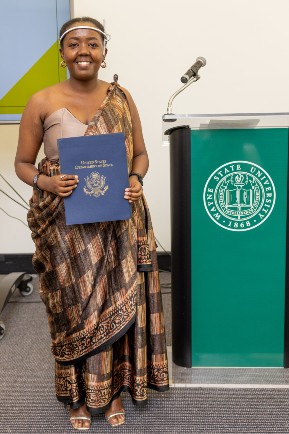 Woman in traditional dress holds a diploma next to a lectern