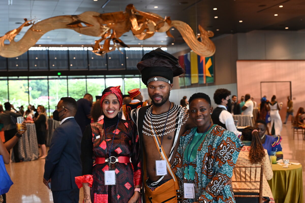 3 Fellows in national dress pose for a group photo in an open foyer. People sit and stand at cocktail tables behind them.