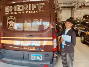 Young woman in a suit points to the letters "CSI" on the back of a van