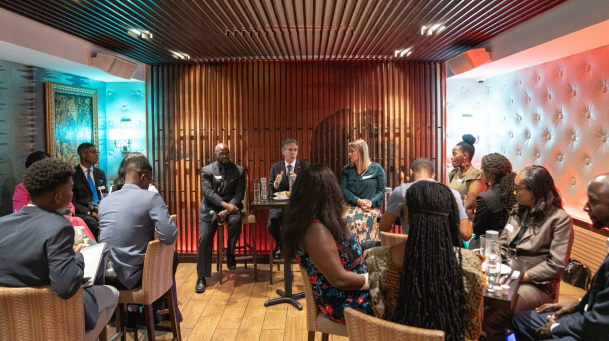 People sit at a high table at the end of the room, the man in the middle holds a microphone