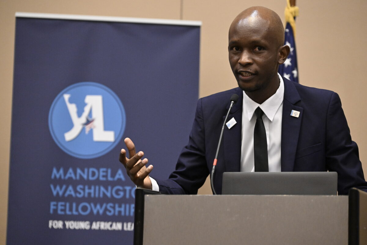 A young man in a suit and tie speaks at a podium, gesturing