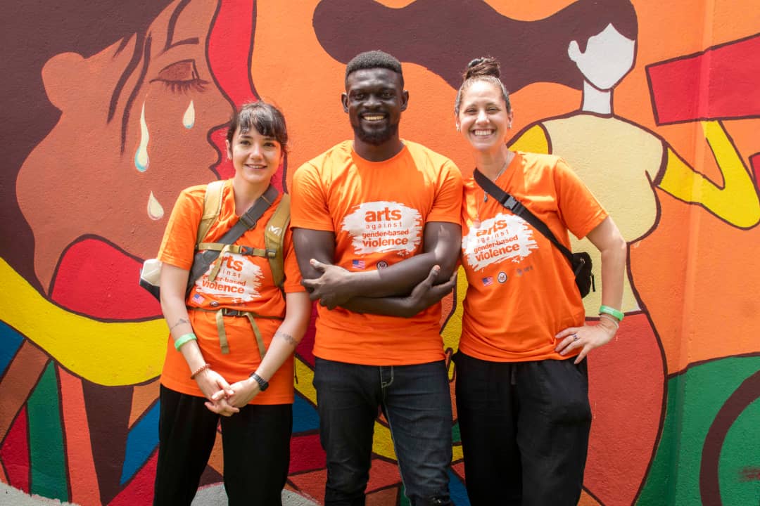 2 women and 1 man pose for a photo in front of a mural; they wear matching orange shirts that say "arts against gender-based violence"