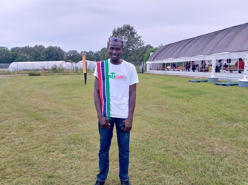 A man in an open field smiles for the camera; he is wearing a white shirt that says “YALI Learns” and has a Gambian flag scarf draped over his shoulder.