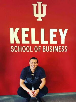A man in a polo shirt crouches in front of a sign that says "Kelley School of Business"