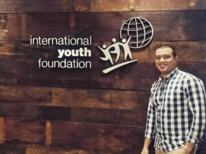 A man in a plaid shirt poses in front of a shiplap wall with a logo for the International Youth Foundation on it