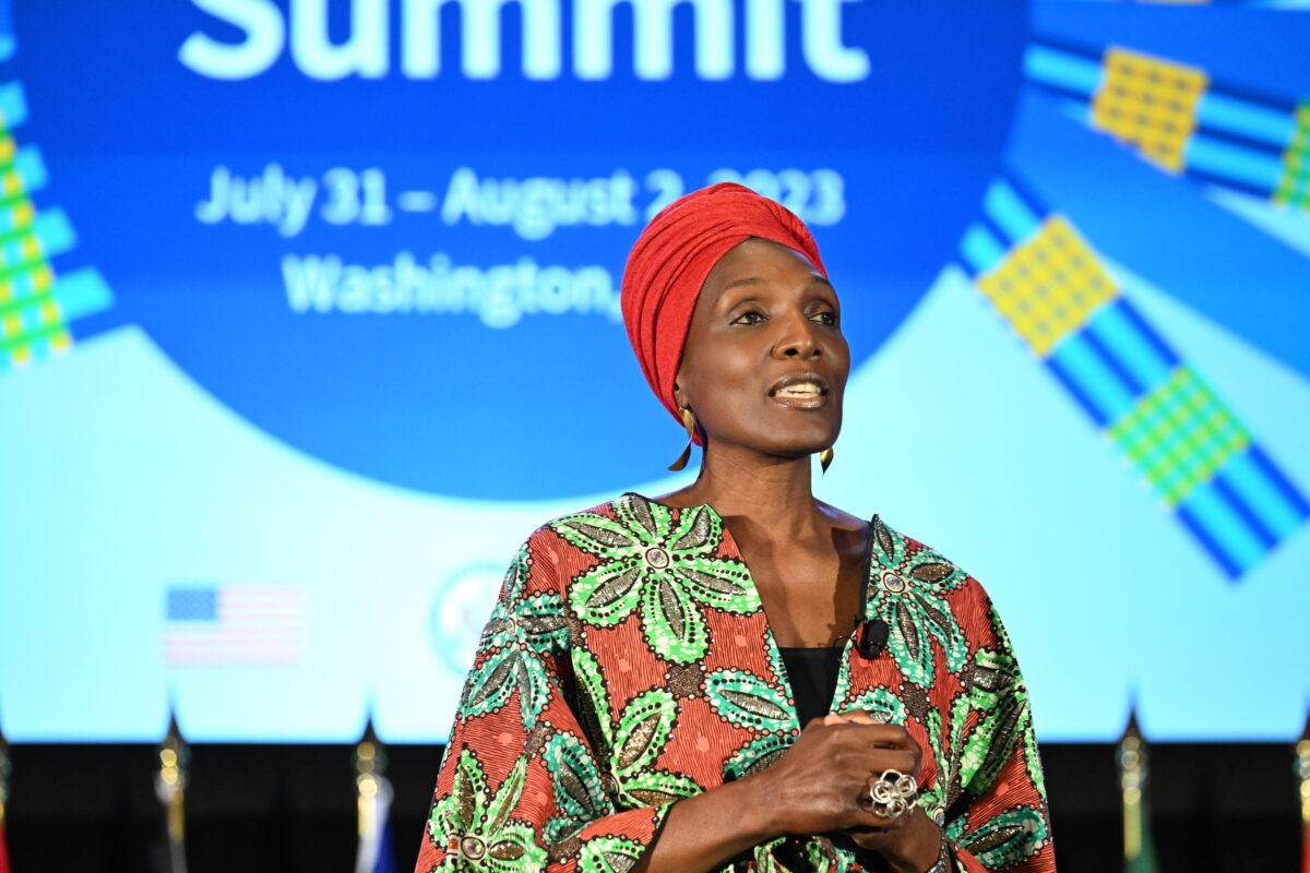 A woman in a red headscarf and red and green large flowered jacket speaks on a stage