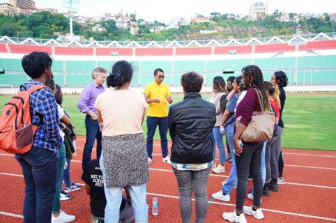 A man in a yellow shirt and sunglasses addresses a group looking at him on the track of a stadium